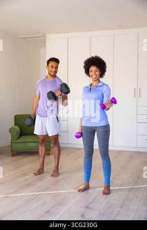Diverse couple in sportswear lifting black and purple dumbbells by armchair in living room Stock Photo