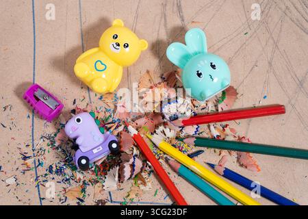 Colorful pencil shavings surround fun pencil sharpeners shaped like a bear, bunny, and dinosaur, resting on a cardboard surface with colored pencils Stock Photo