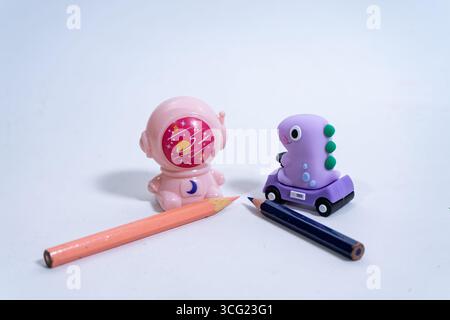Pink astronaut pencil sharpener and purple dinosaur eraser placed near two pencils on a white background, creating a playful and colorful scene Stock Photo