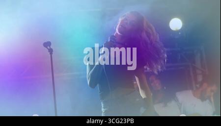 Performing female singer holding handheld microphone on concert stage, with fog and electric guitar Stock Photo