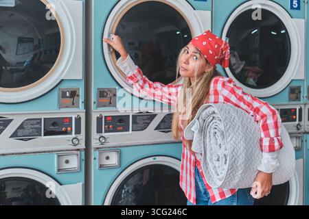 Woman holding blanket and opening dryer door in a laundromat, doing laundry Stock Photo