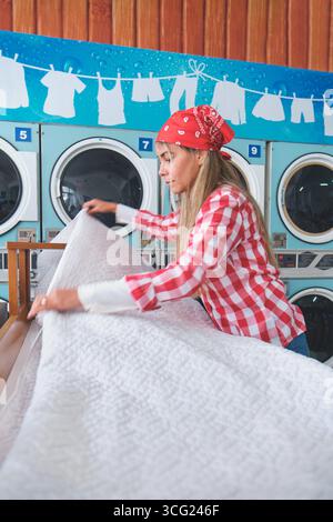 Laundromat worker folding a clean white blanket after a laundry routine, with industrial washing machines in the background Stock Photo