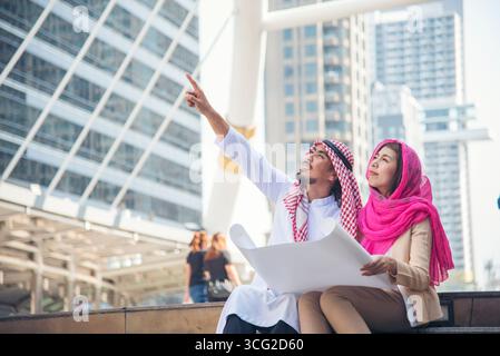 Arab civil construction engineer team consulting project hold blueprint pointing at construction site in muslim UAE city. Diversity teamwork manager p Stock Photo