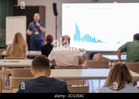 Audience listening to a speaker analyzing data on chart during a business seminar. Seminar setting with participants taking notes. Stock Photo