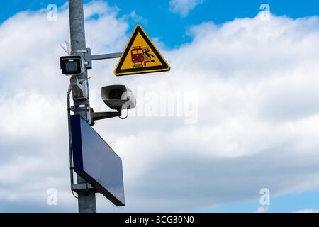 A caution sign signals oncoming trains, while a security camera and speaker are mounted on a pole. The sky above is filled with clouds, contributing to the atmosphere of vigilance. Stock Photo