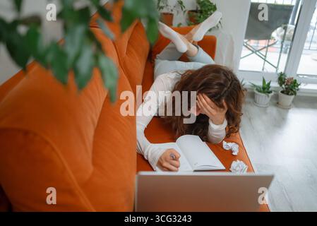 Young woman working from home lying on a sofa using laptop and taking notes on a notebook, she's having a headache and feeling stressed and tired Stock Photo