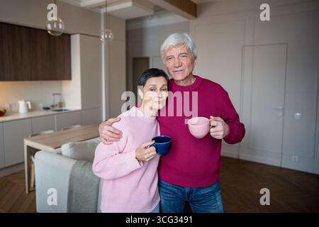 Calm retired wife and husband embracing in morning while drinking tea coffee looking at camera Stock Photo
