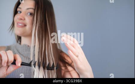 Young woman with hair extensions gently combing hair using comfortable safe comb in beauty salon Stock Photo