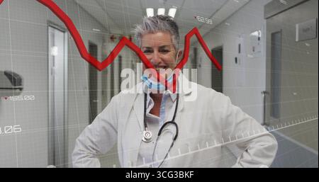 Smiling senior female doctor wearing lab coat and stethoscope in hospital corridor, with red graph Stock Photo