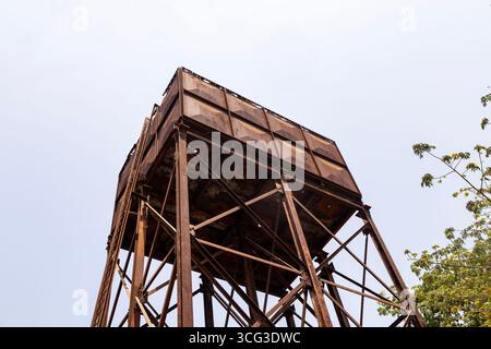 This image features a tall, rusty water tank elevated on metal stilts, set against a clear sky and tree branches, Stock Photo