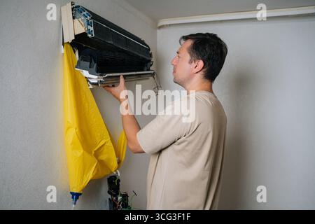Side view of technician disassembling and repairing wall-mounted air conditioner unit, using yellow drainage bag to collect water while working on Stock Photo