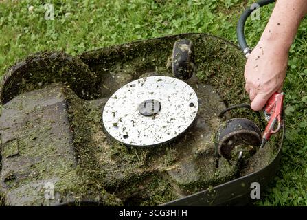 Close-up view of a man's hand blowing off a layer of wet grass stuck under an automated lawnmower using an air compressor — maintenance concept. Stock Photo