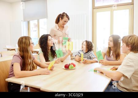 A group of students interact with their teacher in a bright cafeteria, holding colorful cards with educational designs while seated around a table of Stock Photo