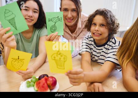 A group of kids and their teacher showcasing hand-drawn food cards during an educational activity. Fresh fruits decorate the table, creating a fun lea Stock Photo
