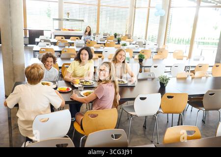 Students are seated at a table in a school cafeteria, enjoying meals together. The room is bright and welcoming, featuring modern furniture and natura Stock Photo