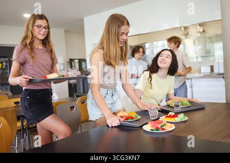 Group of students in a school cafeteria engaging in conversation and enjoying a nutritious meal with fresh food on trays. Bright environment fostering Stock Photo