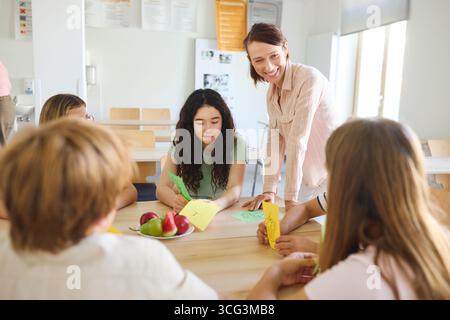 A teacher engaged with students participating in an educational craft activity in a school cafeteria. Fresh produce is placed on the table, fostering Stock Photo