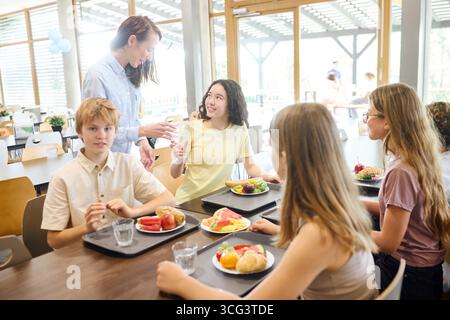 Students in a cafeteria sharing a meal, with a teacher interacting nearby, showcasing diversity. Stock Photo