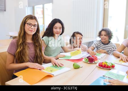 Group of diverse students in a classroom setting exploring nutrition education with fresh fruits and vegetables displayed, showcasing a positive, heal Stock Photo