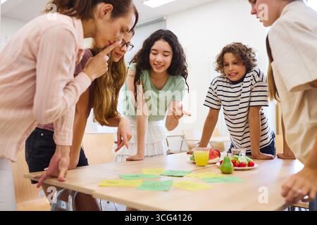 Students of diverse ages discussing and collaborating around a table in a cafeteria, engaging in teamwork and brainstorming ideas, with fresh fruits a Stock Photo