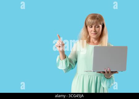 Beautiful mature woman with laptop pointing at something on blue background Stock Photo