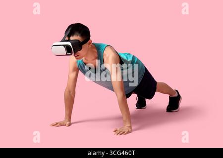 Sporty young Asian man in VR glasses doing push-ups on pink background Stock Photo
