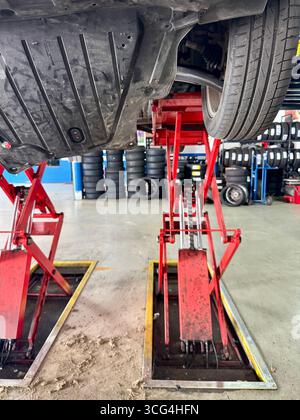 Undercarriage view of a car elevated on red hydraulic lifts inside an automotive repair garage. Stock Photo