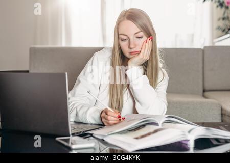 Distance learning. Tired sad girl student sits at home at a table with a laptop and teaches English lessons remotely in quarantine Stock Photo