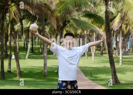 cheerful Asian young man raising coconut joyfully in palm trees grove in Sanya, Hainan, China Stock Photo