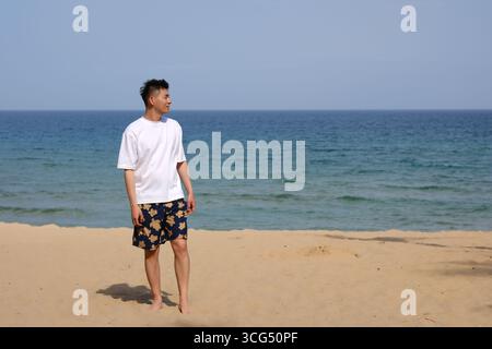 Asian man standing on a sandy beach, looking at blue ocean and sky in Sanya, Hainan, China Stock Photo