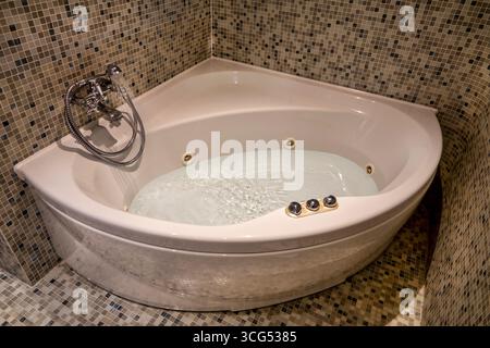 modern hotel bathroom featuring a corner jacuzzi tub with bubbling water. The bathtub is surrounded by elegant mosaic tiles, creating a refined and re Stock Photo
