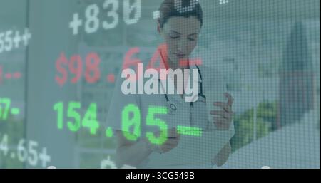 Standing nurse reading clipboard and using smartphone in clinic, with stethoscope and stock ticker Stock Photo