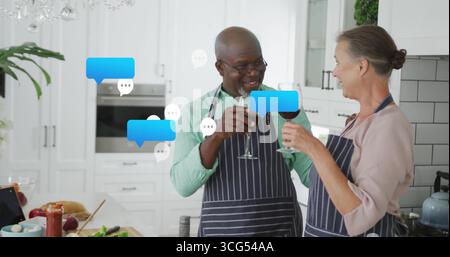 Clinking wine glasses, senior couple in aprons toasting at kitchen island, with digital tablet Stock Photo