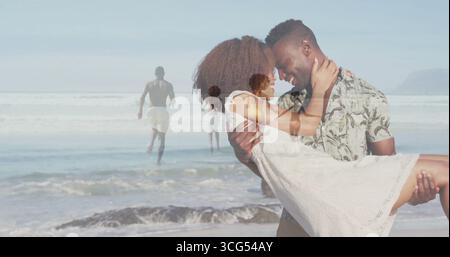 Lifting man wearing floral shirt carrying woman in white sundress at shoreline, with ocean waves Stock Photo