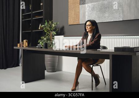 A business professional typing a report on her laptop while seated at a sleek desk in a contemporary office Stock Photo