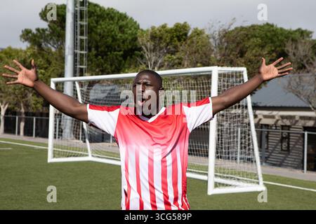 African American man shouting in celebration on artificial turf by soccer goal in red jersey Stock Photo