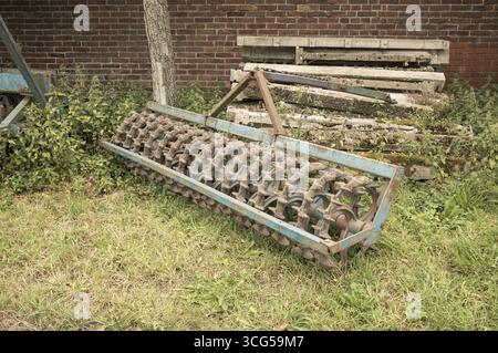 Old cambridge roller laying on grass near a pile of concrete beams and a brick wall Stock Photo