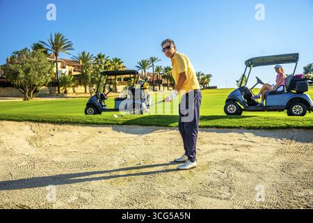 Golfer focuses on a bunker shot while others relax in golf carts on a bright, sunny day at a lush golf course with palm trees Stock Photo