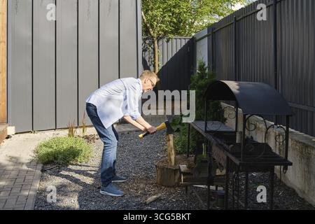 A middle-aged man is chopping a piece of wood in a well-maintained backyard garden. He is dressed casually in a light blue shirt and jeans while surro Stock Photo