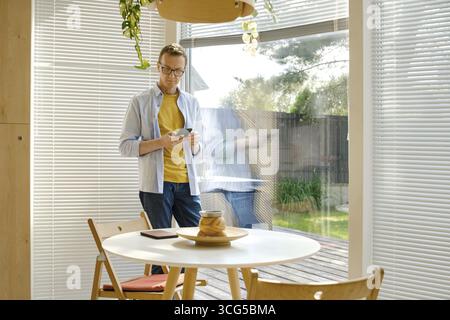 A middle-aged man stands in a bright, modern kitchen checking weather forecast at his smartphone. Sunlight filters through the blinds, illuminating th Stock Photo