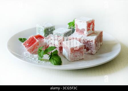 Turkish delight. Lokum, colorful, flavored gelatinous sweets covered in powdered sugar served in a plate on a white table. Mint leaves decoration. Stock Photo