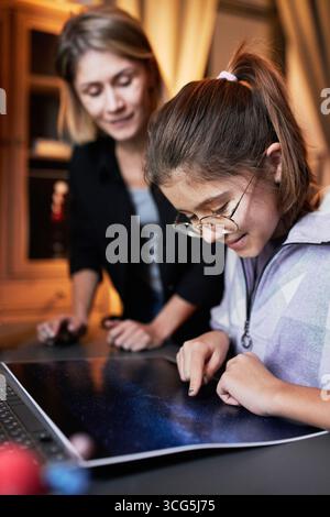 Vertical shot of Caucasian woman helping her joyful daughter with astronomy homework in evening at home Stock Photo