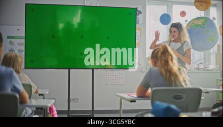 Female Teacher Educating Elementary School Students During Lesson in Modern Classroom. Large Digital Screen with Chroma Key. Green Screen Template for Educational Content or Interactive Visual Effects Stock Photo