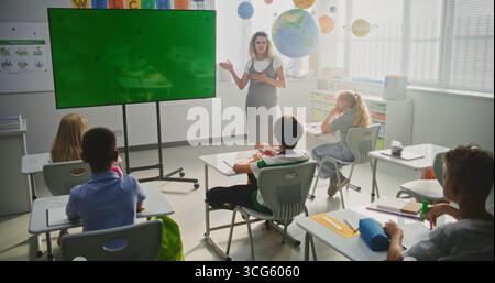Female Teacher Educating Elementary School Students During Lesson in Classroom. Large Digital Screen with Chroma Key. Green Screen Template for Educational Content or Interactive Visuals. Dolly Shot. Stock Photo