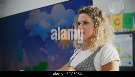 Female Teacher Explaining Basic Information About Weather Factors to Elementary School Students in Modern Classroom. Teacher Educating Geography Using Digital Screen, Talking About Water Cycles. Stock Photo