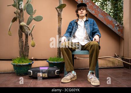 Young man in retro outfit with cassette player and cactus pots sitting in urban backyard. Vintage fashion, youth subculture, 90s nostalgia, indie lifestyle, analog music era. Stock Photo