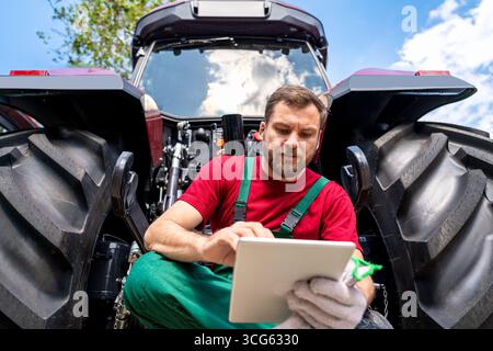 Low angle portrait of farmer using digital tablet while sitting near tractor in rural area. Stock Photo