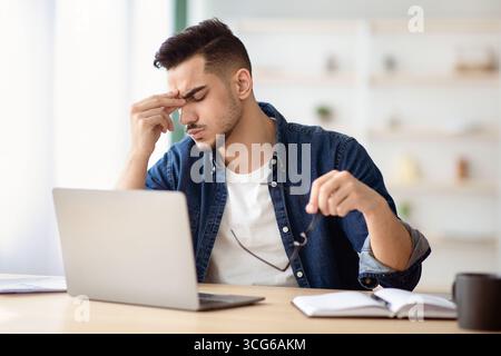 Exhausted arab guy sitting at workdesk, removing glasses Stock Photo