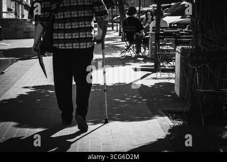 A powerful black-and-white street photograph showing a man walking with the aid of a cane along a sunlit sidewalk. Captured from behind, the image hig Stock Photo