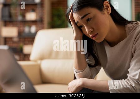 Closeup of unhappy freelancer working from home, reading emails with financial issues on laptop. Asian female manager sadly looking at device screen, feeling tired and overworked, sitting in apartment Stock Photo